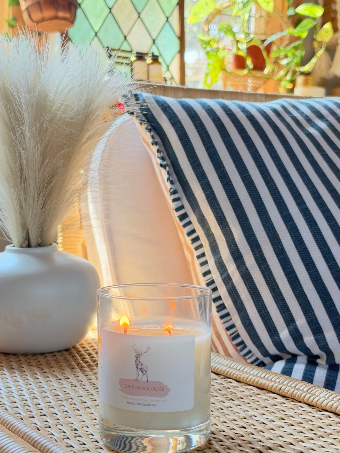 Driftwood Rose double wick candle burning beside a striped pillow and vase